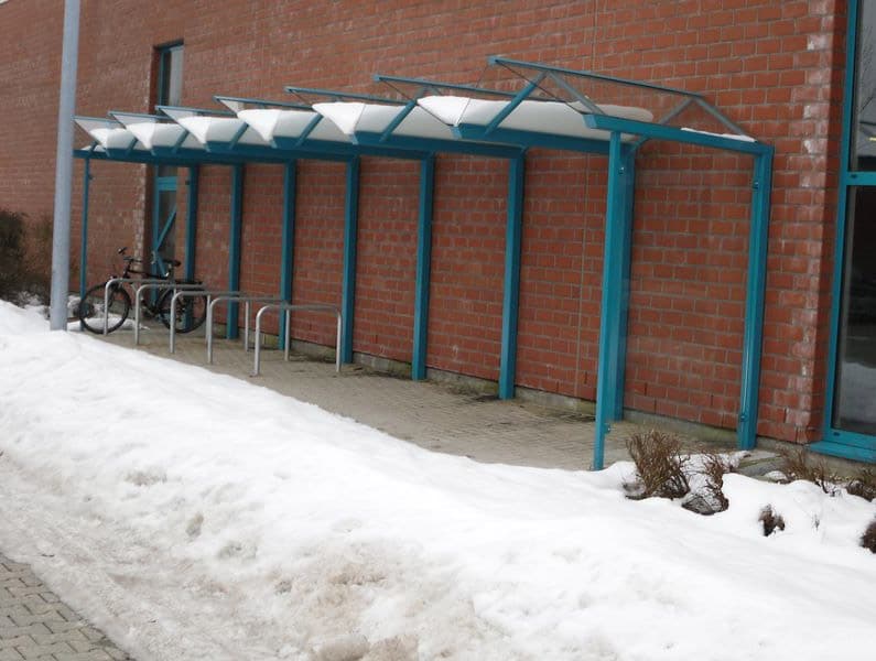 Eine Fahrradüberdachung aus Stahl, auf der sich Schnee angesammelt hat, vor einer Steinmauer